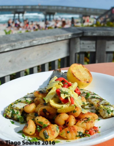 Seasoned grilled fish paired with fresh scallops, topped with roasted vegetables and lemon displayed on a round white plate. The background is a beach pier on a sunny summer day in Florida. Photographed by Tiago with the Shots.