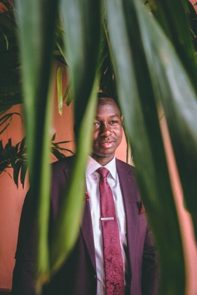 Gentleman in burgandy suit with his hands in his pants pockets. He is standing behind a palm tree in front of a salmon colored building- while looking off into the distance. Photographed by Tiago with the Shots.