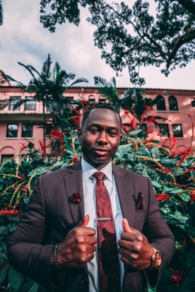 Man in a burgandy suit , accompanied by a red rose attached to his blazer and burgandy handkerchiefs located in his left blazer pocket. The gentleman is looking directly a the Tiago the photgrapher, while holding his blazer open with both hands. Photographed by Tiago with the Shots.