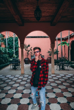 Young man holding his chin with one hand in his pocket. He is standing in an open and closed building with skinny pillars and alor of greenery. Photographed by Tiago with the Shots.